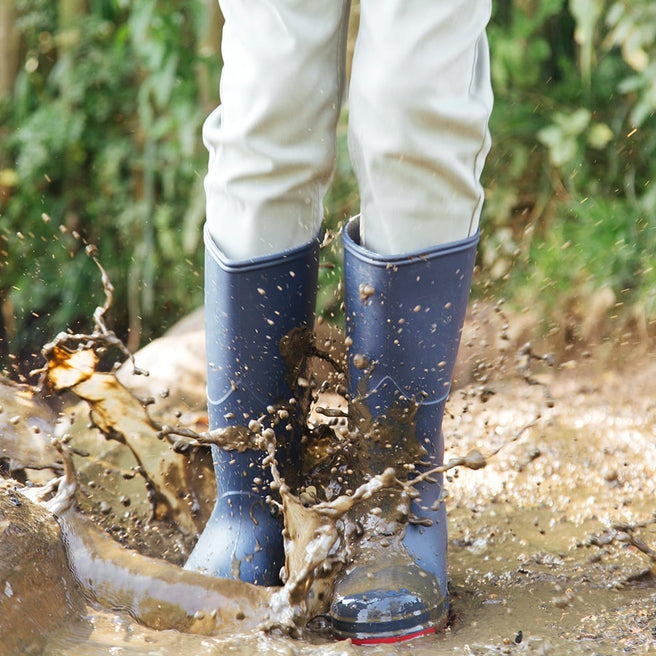 A person in 'Classic Wellies Navy' stands in a muddy puddle, creating a splash of mud with their boots. Green foliage in the background.